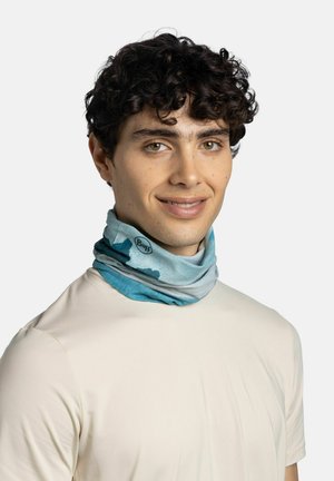 Joven con cabello rizado que lleva un braga de cuello Buff azul con estampado y una camisa beige claro, sonriendo ligeramente.