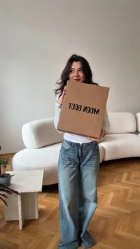 Scatola di cartone con la scritta "MOON BOOT" stampata in nero, tenuta da una persona. Sullo sfondo un divano curvo di colore chiaro e un piccolo tavolo con una pianta.