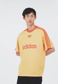 Yellow athletic shirt with red accents, featuring the Adidas logo and three-striped sleeves. Lightweight fabric with a relaxed fit.