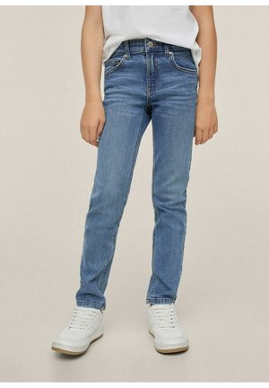 Enfant portant un jean skinny bleu clair avec des baskets blanches et un t-shirt blanc, debout devant un fond uni.