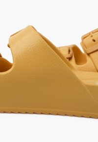 Yellow molded sandals with a textured surface, pronounced heel, and adjustable strap. Features a zigzag sole for traction.