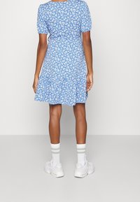 Donna che indossa un vestito blu a fiori di lunghezza al ginocchio con maniche a sbuffo, calzini bianchi con strisce verdi e sneaker bianche, in piedi contro uno sfondo semplice.