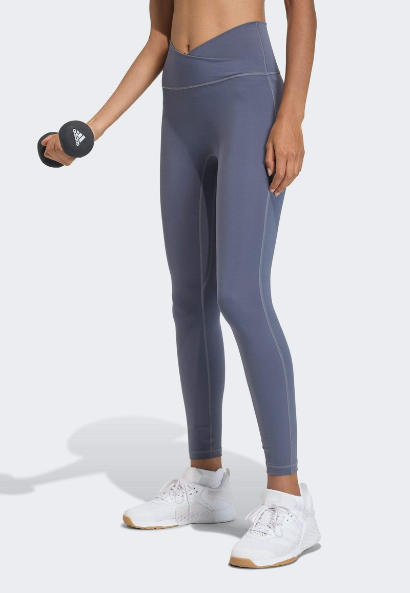 Grijze legging met hoge taille en contrasterende naden, gecombineerd met witte sportschoenen. Het model houdt een zwarte halter in één hand vast.