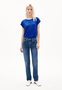 Blauwe T-shirt met korte mouwen, relaxte fit, gecombineerd met lichtblauwe recht gesneden jeans. Model draagt witte schoenen, voorgrond tegen een eenvoudige witte achtergrond.