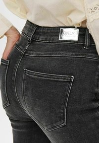 Black denim jeans with a high waist, featuring a shiny silver label, subtle stitching, and two back pockets for detail.