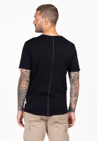 Camiseta negra de manga corta con dobladillo sin terminar y una costura trasera prominente. La tela parece suave, complementando unos pantalones cortos de carga beige.