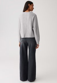 Donna in un top manica lunga grigio chiaro e jeans neri a vita alta e a gamba larga, in piedi di spalle contro uno sfondo neutro.