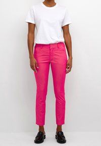 Pantalon rose slim en denim, associé à un t-shirt blanc. Le pantalon présente une texture lisse et des poches classiques. Mocassins noirs portés.