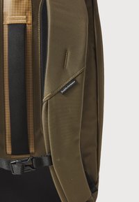 Dark olive green backpack with textured fabric, black straps, and a Peak Performance label. Features a grid-patterned pocket. Adjustable buckle.