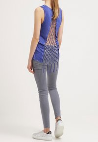 Top sin mangas azul con una gran espalda descubierta y un diseño entrecruzado, con flecos en la parte inferior, combinado con jeans grises elásticos y zapatillas blancas.
