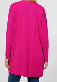 Tunique à manches longues de couleur rose vif, présentant une coupe décontractée, des détails côtelés aux épaules et des fentes latérales pour faciliter les mouvements.