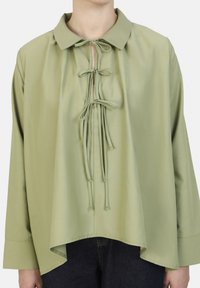 Blusa larga de manga larga, color verde claro, con cuello, detalle de lazo en el escote y dobladillo curvado. Tela suave con una textura ligeramente brillante.