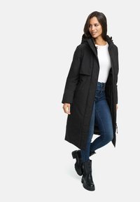 Black long parka with a hood, two front pockets, and zipper detail. Worn over a white top and blue jeans, paired with black chunky boots.