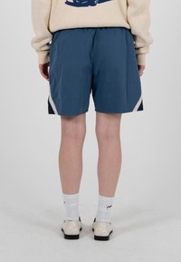 Person trägt blaue Shorts mit hellen, schrägen Streifen, cremefarbenen Pullover, weiße Socken und cremefarbene Schuhe und steht vor einem einfarbigen hellen Hintergrund.