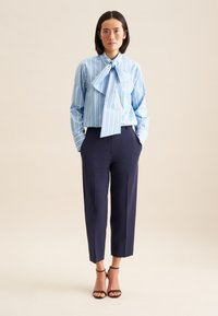 Camicia a righe blu e bianche con un grande colletto a fiocco, abbinata a pantaloni a vita alta blu navy e tacchi neri con cinturini.
