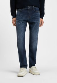 Jeans de mezclilla azul oscuro con un corte clásico, diseño de cinco bolsillos y detalles sutiles de desgaste. Llevados con zapatillas de color crema.