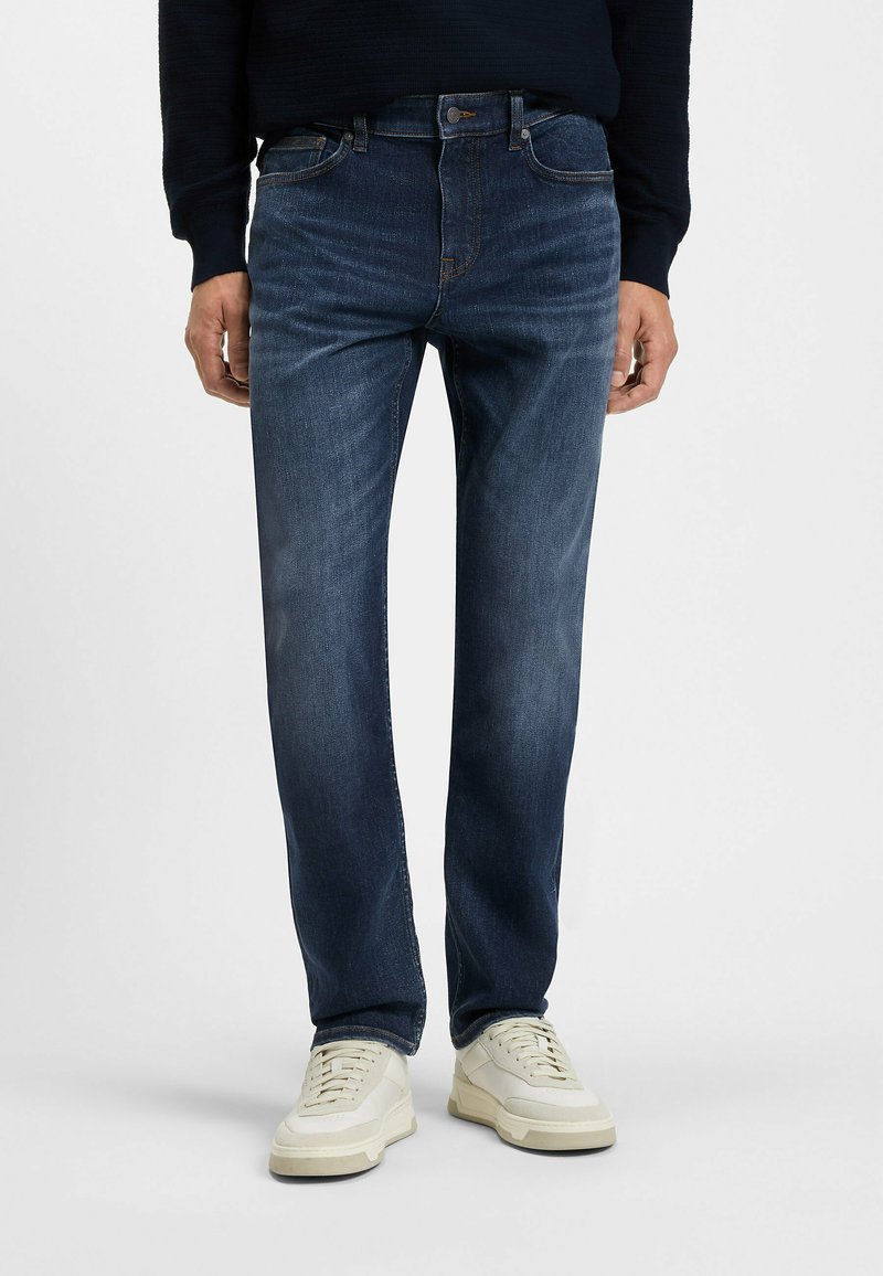 Jeans de mezclilla azul oscuro con un corte clásico, diseño de cinco bolsillos y detalles sutiles de desgaste. Llevados con zapatillas de color crema.