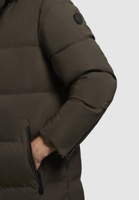 Olijfgroene pufferjack met een matte afwerking, voorzien van een ritszak op de mouw en een gewatteerde textuur voor isolatie.