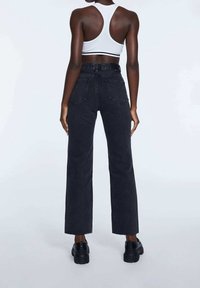 Persona vestida con jeans negros de cintura alta, top corto blanco de tirantes y zapatos chunky negros, de pie con la espalda hacia el frente sobre un fondo liso.