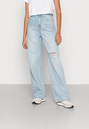 Jean large bleu clair avec taille haute et détail déchiré au genou, fabriqué en tissu denim, associé à des baskets blanches.