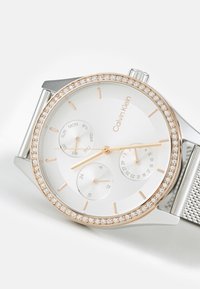Reloj redondo de plata con una esfera blanca, acentos en tono oro rosa y un bisel con cristales brillantes. Correa de malla y tres sub-esferas que indican los días y la fecha.