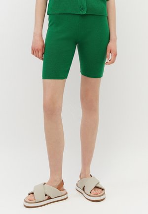 Persoon draagt groene gebreide shorts en bijpassend topje, gecombineerd met beige sandalen met kruisbandjes en witte zolen, staand op een witte achtergrond.
