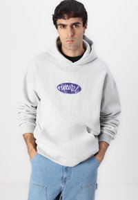 Sweatshirt gris avec une poche avant, arborant un logo brodé violet. Le design a une coupe décontractée et des poignets côtelés. Porté avec un jean bleu clair.