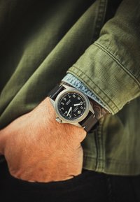 Reloj negro con caja de acero inoxidable, números blancos y correa de cuero negra, llevado en una mano que descansa en un bolsillo de chaqueta verde.