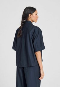 Camicetta blu navy a maniche corte con colletto, realizzata in tessuto leggero. Caratterizzata da una vestibilità ampia, fessure laterali e una piega posteriore per facilitare il movimento.