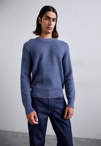 Suéter azul de punto con puños y dobladillo acanalados, combinado con jeans de mezclilla oscuros. Textura suave, diseño de cuello redondo y color sólido.