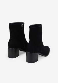 Botas de tobillo de ante negro con puntera redondeada, tacón bloque de altura media y cremallera lateral. Presenta tachuelas plateadas a lo largo del tacón como detalle.