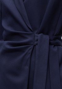 Σκούρα μπλε υφασμάτινη ζώνη που σφίγγει ένα ένδυμα στη μέση, δημιουργώντας μαζεμένες πτυχές και μια λεία, ελαφρώς υφή επιφάνεια.