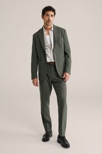 Bărbat îmbrăcat într-un costum verde, cu o cămașă albă și o curea maro, stând cu o mână în buzunar și cu un fundal bej neutru.