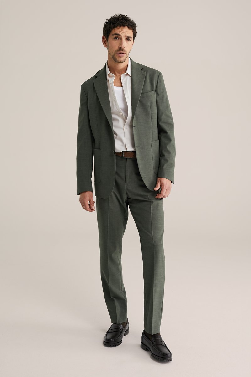 Bărbat îmbrăcat într-un costum verde, cu o cămașă albă și o curea maro, stând cu o mână în buzunar și cu un fundal bej neutru.