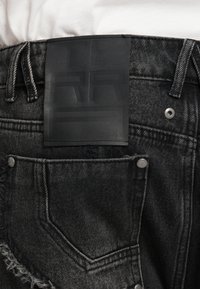 Jeans de ganga preta com um rótulo em couro texturizado na parte de trás, com uma bainha desgastada e detalhes em metal prateado.