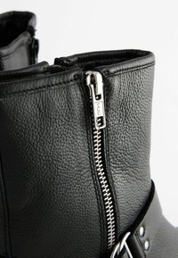 Gros plan d'une botte en cuir noir texturé avec une fermeture éclair YKK argentée et un détail de boucle sur le côté sur fond blanc.