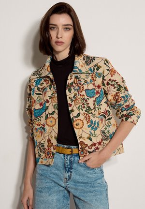 Donna che indossa una giacca beige ricamata con motivi floreali sopra una maglia nera, jeans azzurri e una cintura gialla, in piedi contro una parete semplice.