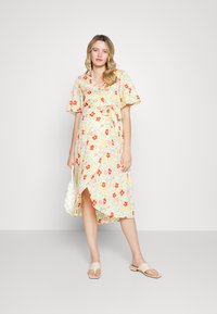 Vestido envolvente floral com mangas curtas, cintura com laço e uma fenda alta. O tecido é leve, com fundo amarelo e padrões em vermelho, rosa e verde.