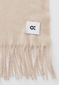 Écharpe beige à franges en matériau doux avec une finition texturée. Présente une étiquette blanche avec un logo noir.