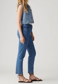 Beskuren denimväst, blå högmidjade jeans och svarta sandaler med remmar. Västen har knappar och rå kant; jeansen har rak passform med ljus tvätt.