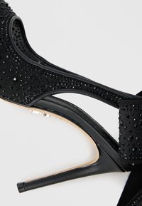 Sandale negre cu toc înalt, având partea superioară texturată decorată cu pietre prețioase negre, vârf deschis și toc subțire stiletto.