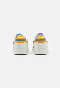 Ett par vita sneakers med blå och gula accenter, sedda från baksidan, med en "diadora" logotyp på hälen.