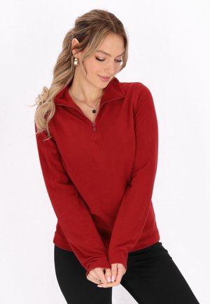 Rode fleece pullover met een half-rits ontwerp, lange mouwen en een zachte textuur. Voorzien van een hoge kraag en een aansluitende silhouette. Zwarte broek eronder gedragen.