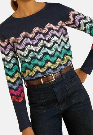 Femme portant un pull bleu marine avec des rayures ondulées multicolores et un jean noir avec une ceinture marron.