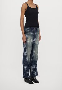 Femme portant un débardeur noir, un jean large en denim bleu à clous, et des bottines noires à bout pointu, se tenant devant un fond blanc.