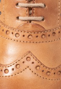 Chaussure en cuir tan avec des détails de brogue décoratifs, des accents de couture contrastants et des perforations rondes le long du bord supérieur et de la zone des orteils.