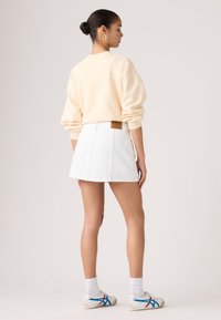 Γυναίκα που στέκεται, φοράει ένα κρεμ φούτερ, λευκή μίνι φούστα, λευκές κάλτσες και λευκά αθλητικά παπούτσια με μπλε και κόκκινες ρίγες, στραμμένη με την πλάτη.