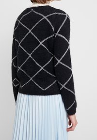 Zwarte zachte trui met een diamantpatroon in witte draad, met lange mouwen en een geribde zoom. Draagt over een lichtblauwe geplooide rok.
