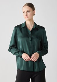 Žalia satino marškiniai su mygtukų uždarymu, ilgomis rankovėmis ir klasikinėmis apykaklėmis. Lygi tekstūra ir šiek tiek laisvas kirpimas.