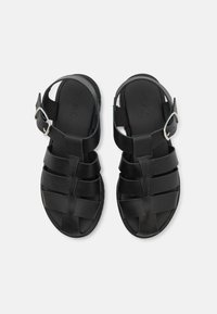 Sandalias de cuero negro con amplias correas en forma de cruz, diseño de punta abierta y una correa en el tobillo que cuenta con una hebilla de metal plateado. Suela plana.